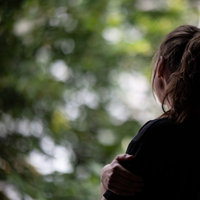 Ein Mädchen mit braunen Haaren, die es zu einem Pferdeschwanz zusammengebunden hat, steht traurig an einem Fenster.