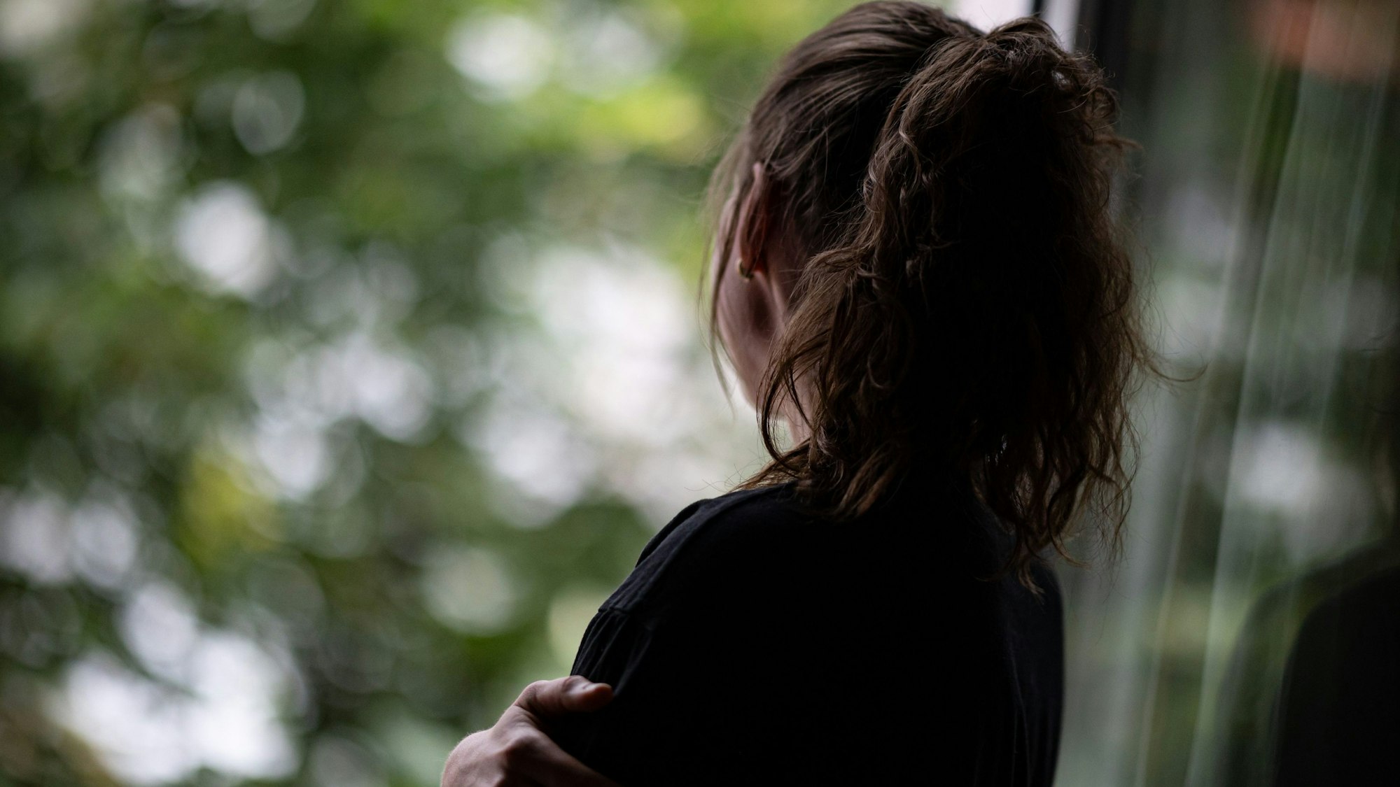 Ein Mädchen mit braunen Haaren, die es zu einem Pferdeschwanz zusammengebunden hat, steht traurig an einem Fenster.