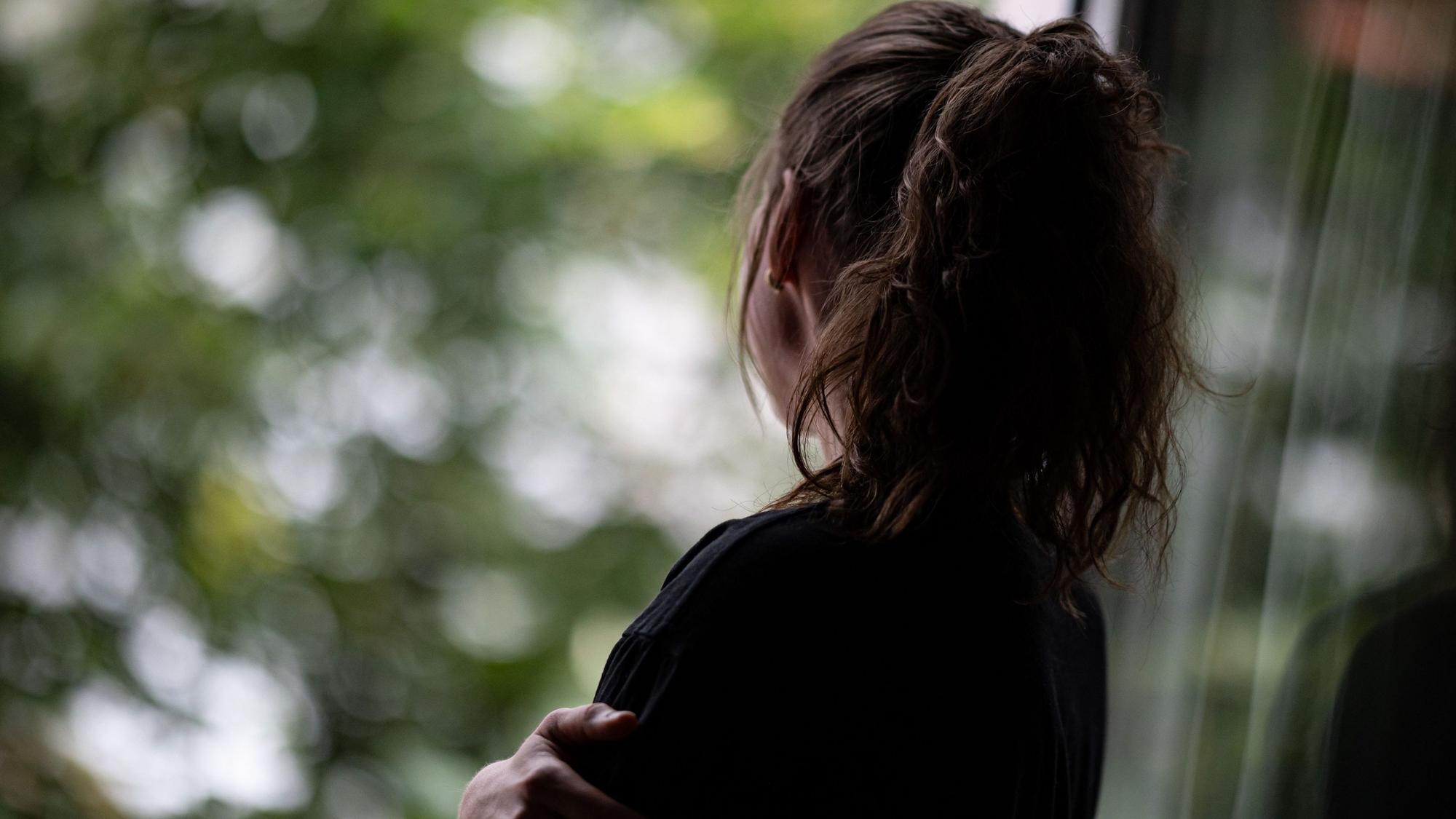 Ein Mädchen mit braunen Haaren, die es zu einem Pferdeschwanz zusammengebunden hat, steht traurig an einem Fenster.