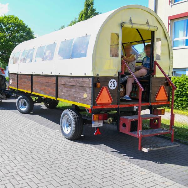 Ein von einem Traktor gezogener Planwagen ist zu sehen. Darin sitzen zwei Menschen in sommerlicher Kleidung.