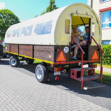 Ein von einem Traktor gezogener Planwagen ist zu sehen. Darin sitzen zwei Menschen in sommerlicher Kleidung.