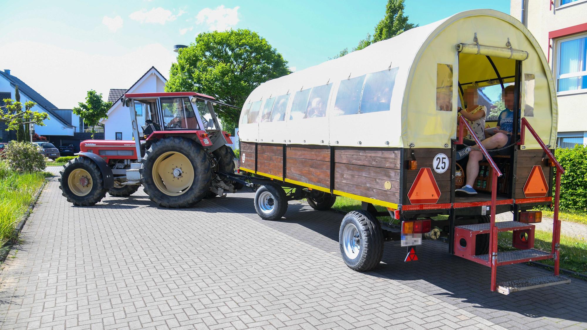 Ein von einem Traktor gezogener Planwagen ist zu sehen. Darin sitzen zwei Menschen in sommerlicher Kleidung.