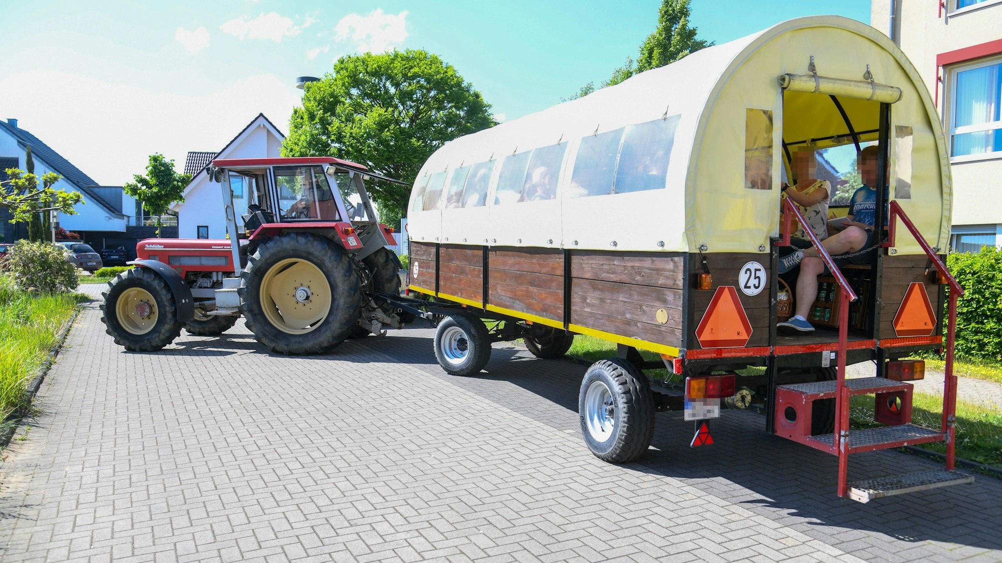 Ein von einem Traktor gezogener Planwagen ist zu sehen. Darin sitzen zwei Menschen in sommerlicher Kleidung.