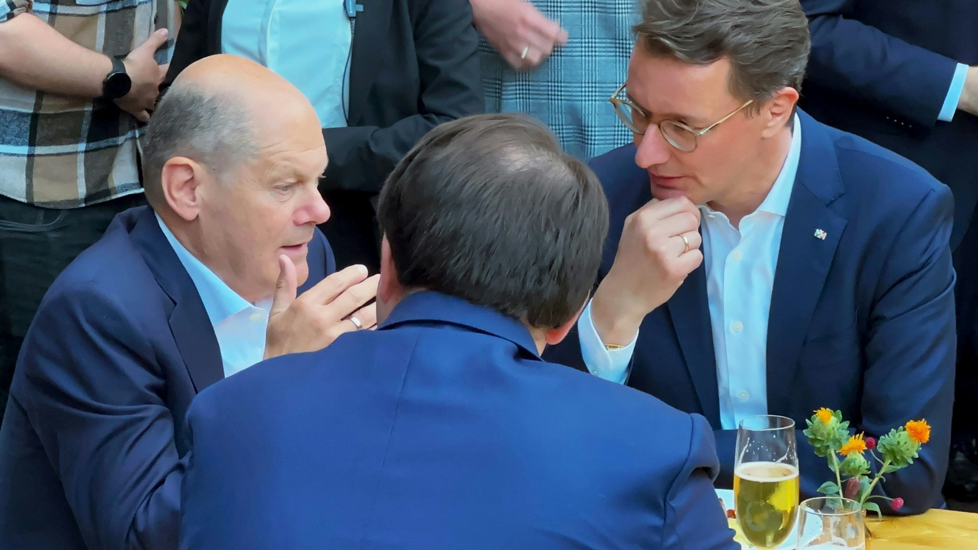 Bundeskanzler Olaf Scholz (SPD) im Gespräch mit Hendrik Wüst und Staatskanzleichef Nathanael Liminski. Sie stehen um einen Biertisch, Scholz spricht, die anderen hören zu.