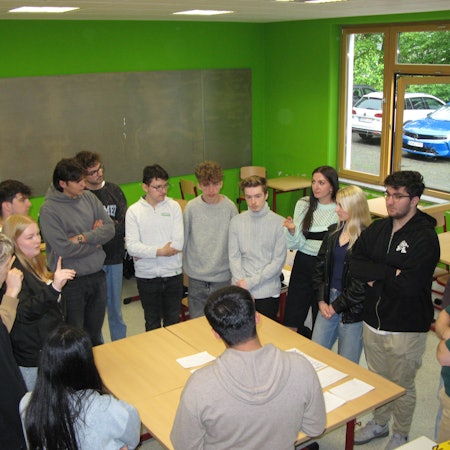 15 jugendliche Schülerinnen und Schüler stehen mit ihrem Geschichtslehrer im Kreis um einen großen Tisch.