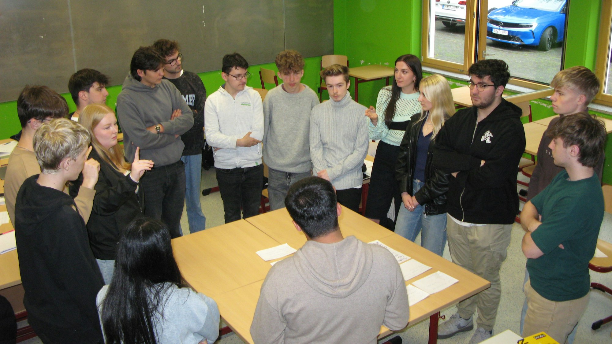 15 jugendliche Schülerinnen und Schüler stehen mit ihrem Geschichtslehrer im Kreis um einen großen Tisch.