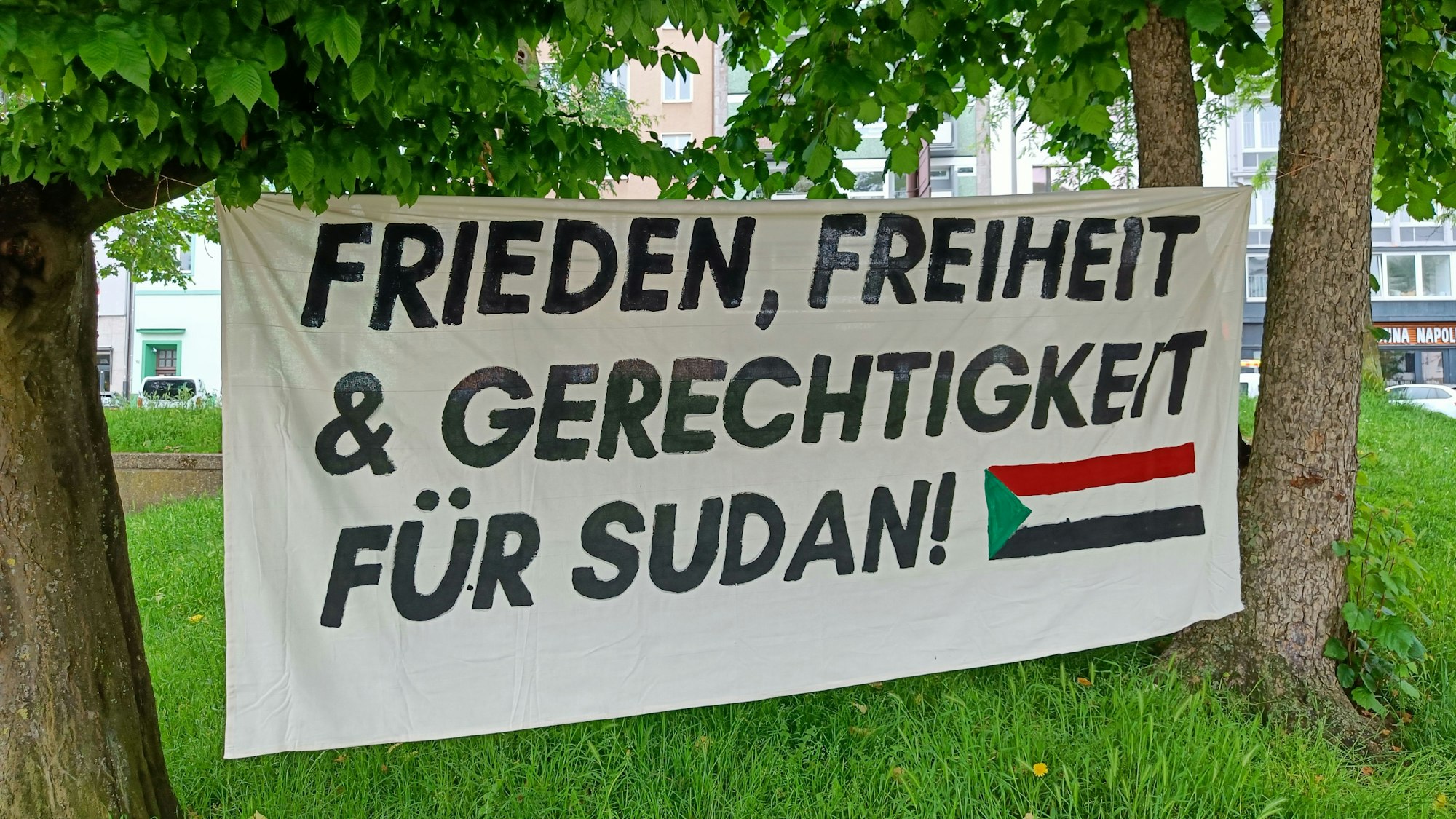 Ein Banner ist zwischen zwei Bäumen gespannt. Darauf steht: „Frieden, Freiheit und Gerechtigkeit für Sudan“.
