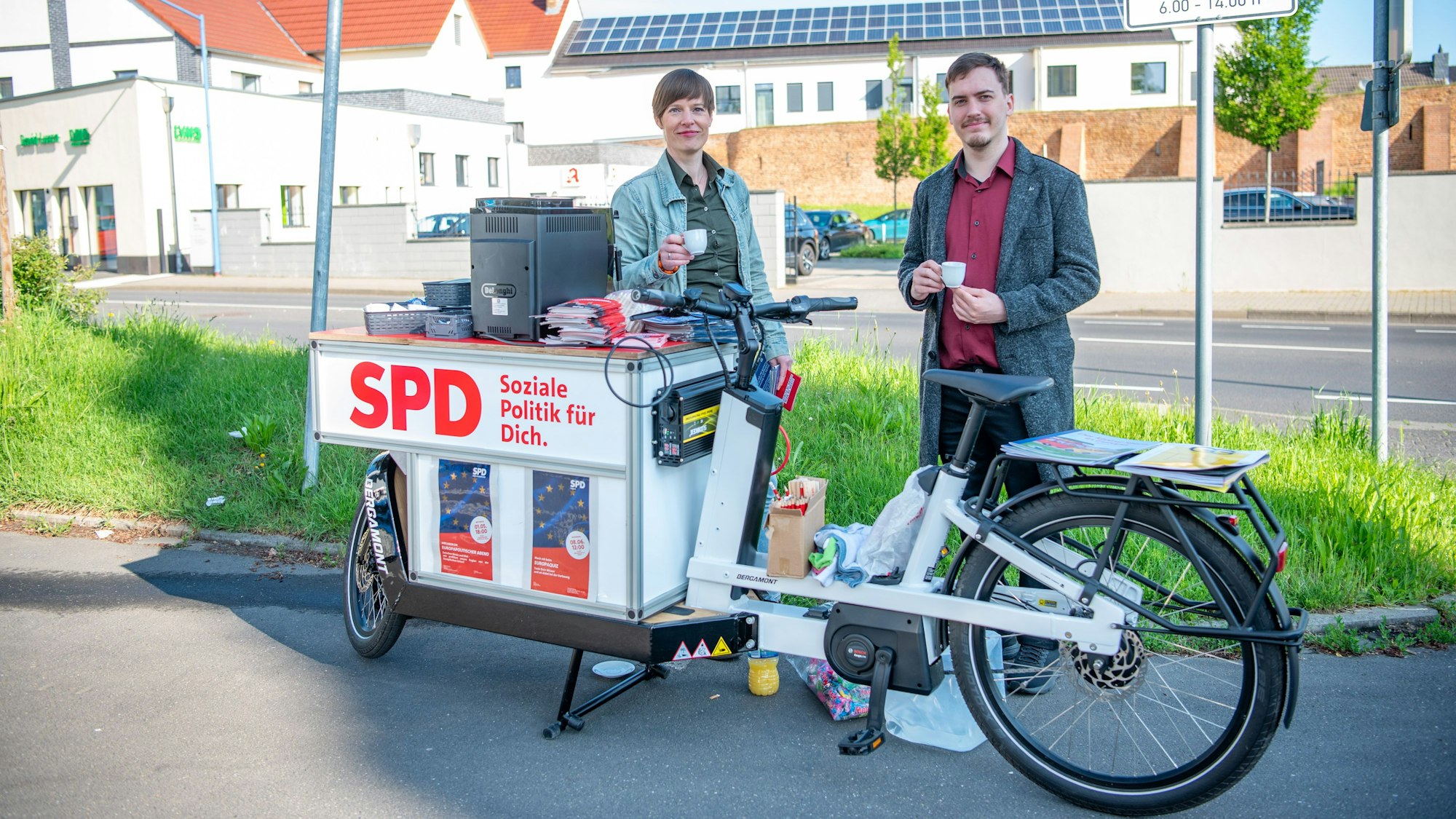 Zwei SPD-Mitglieder stehen mit einer Tasse Espresso an einem weißen Lastenrad, auf dem das Logo der SPD prangt.