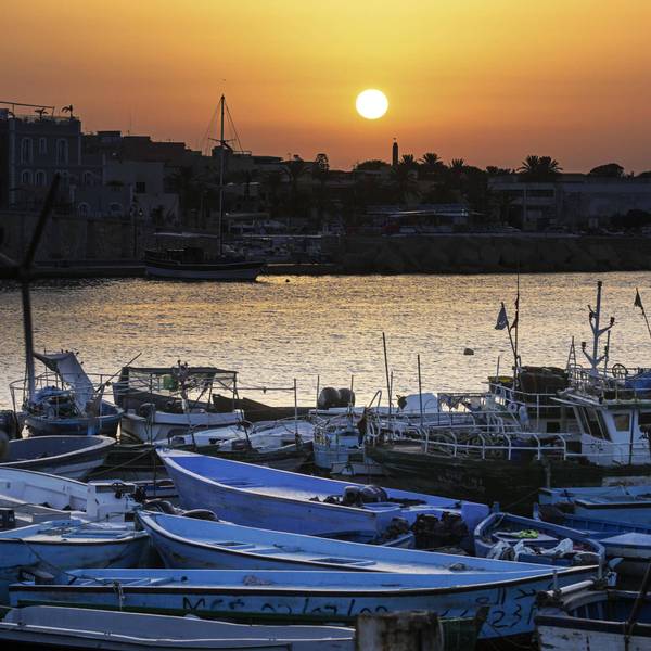 Der Hafen von Lampedusa mit einem„ Bootsfriedhof“: Im Vordergrund Boote, mit denen Geflüchtete die südlichste Insel Italiens erreichen. Die Boote aus dünnen Metallschichten liegen häufig verlassen am Strand, wo die Menschen nach der Flucht ankommen.