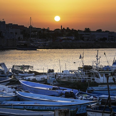 Der Hafen von Lampedusa mit einem„ Bootsfriedhof“: Im Vordergrund Boote, mit denen Geflüchtete die südlichste Insel Italiens erreichen. Die Boote aus dünnen Metallschichten liegen häufig verlassen am Strand, wo die Menschen nach der Flucht ankommen.