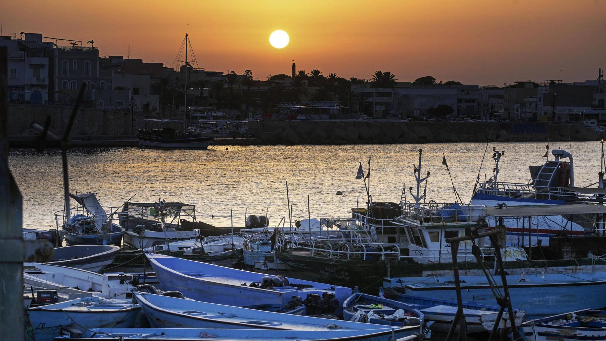 Der Hafen von Lampedusa mit einem„ Bootsfriedhof“: Im Vordergrund Boote, mit denen Geflüchtete die südlichste Insel Italiens erreichen. Die Boote aus dünnen Metallschichten liegen häufig verlassen am Strand, wo die Menschen nach der Flucht ankommen.