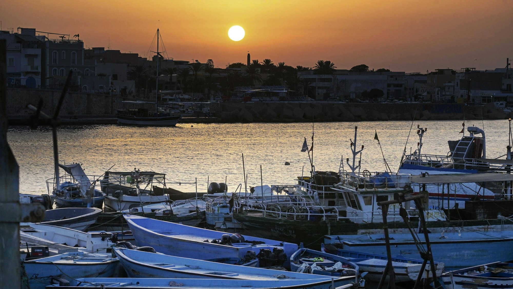 Der Hafen von Lampedusa mit einem„ Bootsfriedhof“: Im Vordergrund Boote, mit denen Geflüchtete die südlichste Insel Italiens erreichen. Die Boote aus dünnen Metallschichten liegen häufig verlassen am Strand, wo die Menschen nach der Flucht ankommen.