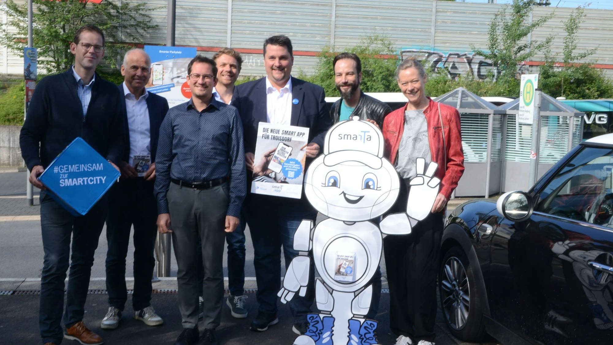 Männer und Frauen stehen auf einem Parkplatz. im Hintergrund ist eine Bushaltestelle zu sehen, vor der Gruppe steht die winkende Pappfigur eines Roboters.