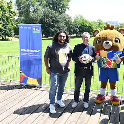 Vier Männer stehen mit einem Maskottchen vor der Wiese des Rheinpark