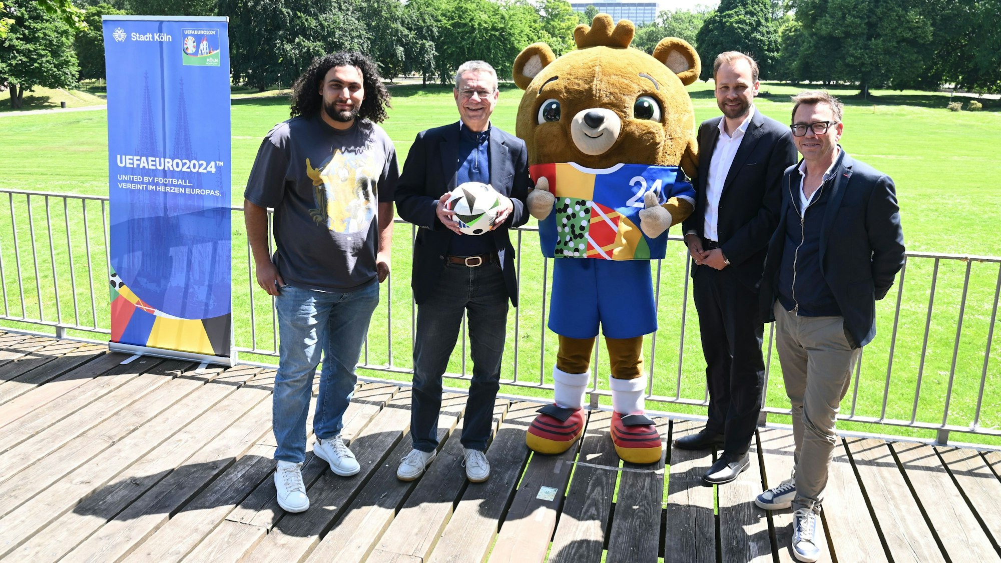 Vier Männer stehen mit einem Maskottchen vor der Wiese des Rheinpark