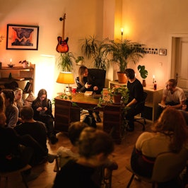 Zu sehen sind Menschen in einem schwach beleuchteten Wohnraum, die auf Stühlen sitzen.