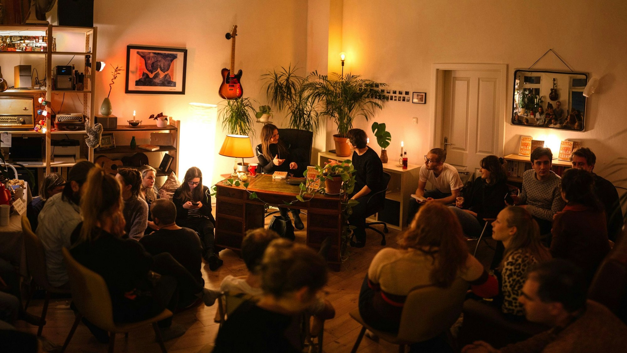 Zu sehen sind Menschen in einem schwach beleuchteten Wohnraum, die auf Stühlen sitzen.