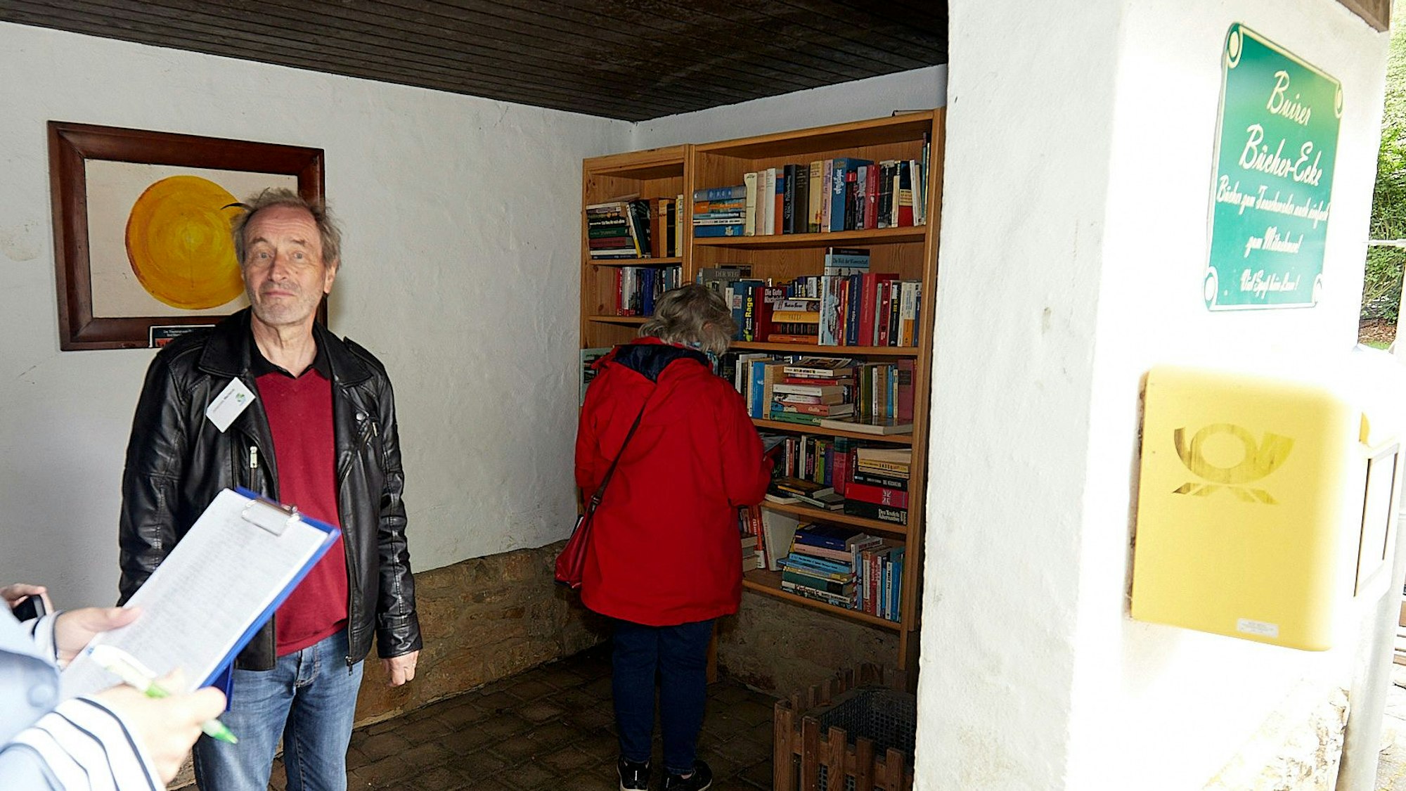 Ein Mann steht im Buswartehäuschen in Nettersheim-Buir. Im Hintergrund ist eine Frau zu erkennen, die an einem Regal steht, das mit zahlreichen Büchern gefüllt ist.