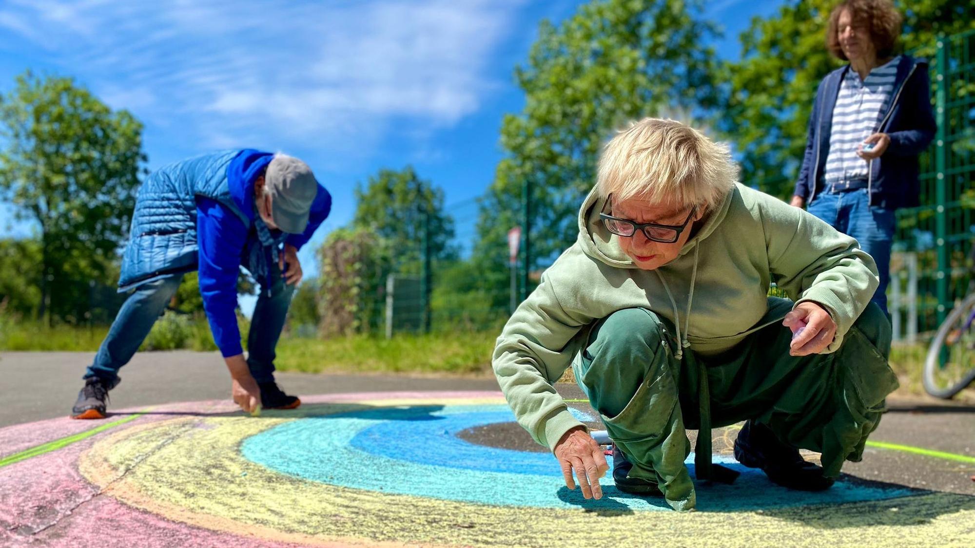 Anne Bergmann, Leo Schmidt und Thomas Latzke malen mit Straßenkreide einen Regenbogen auf die Straße.