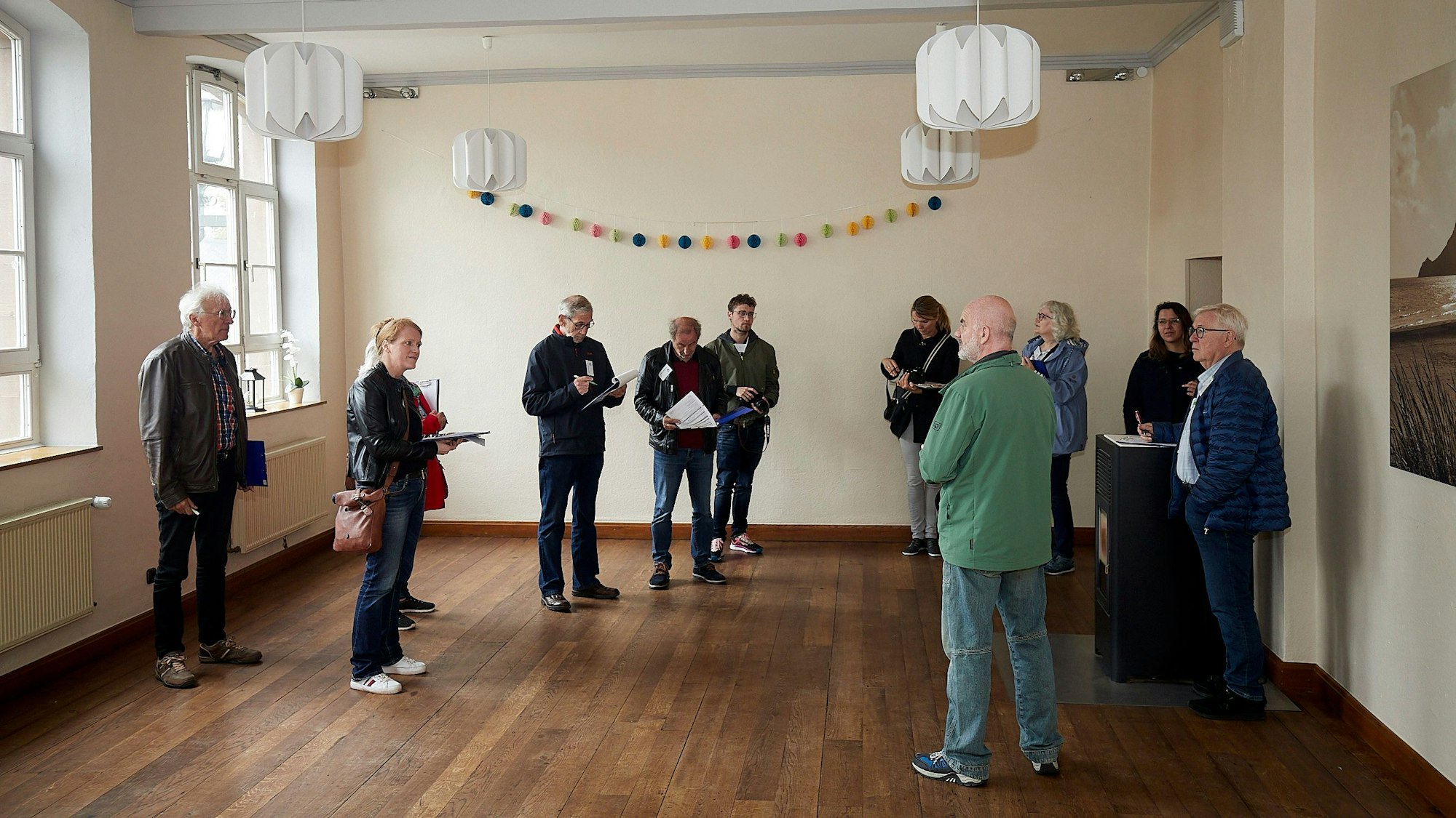 Einige Frauen und Männer stehen in einem kleinen Saal mit braunem Parkettboden und hellen Wänden.