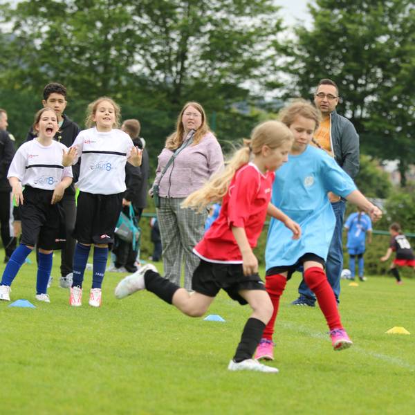 Zwei Mädchen spielen Fußball, Zuschauer stehen am Rand und fiebern mit.