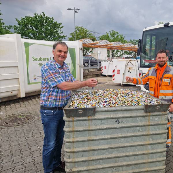 Zu sehen sind zwei Mitarbeiter vom Stadtservice, die an einem mit Kronkorken gefüllten Behältnis stehen.