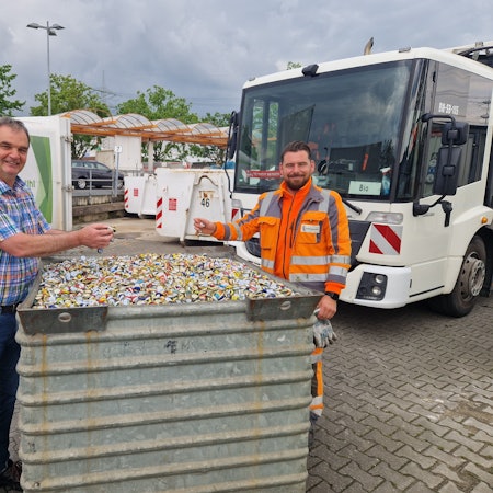 Zu sehen sind zwei Mitarbeiter vom Stadtservice, die an einem mit Kronkorken gefüllten Behältnis stehen.