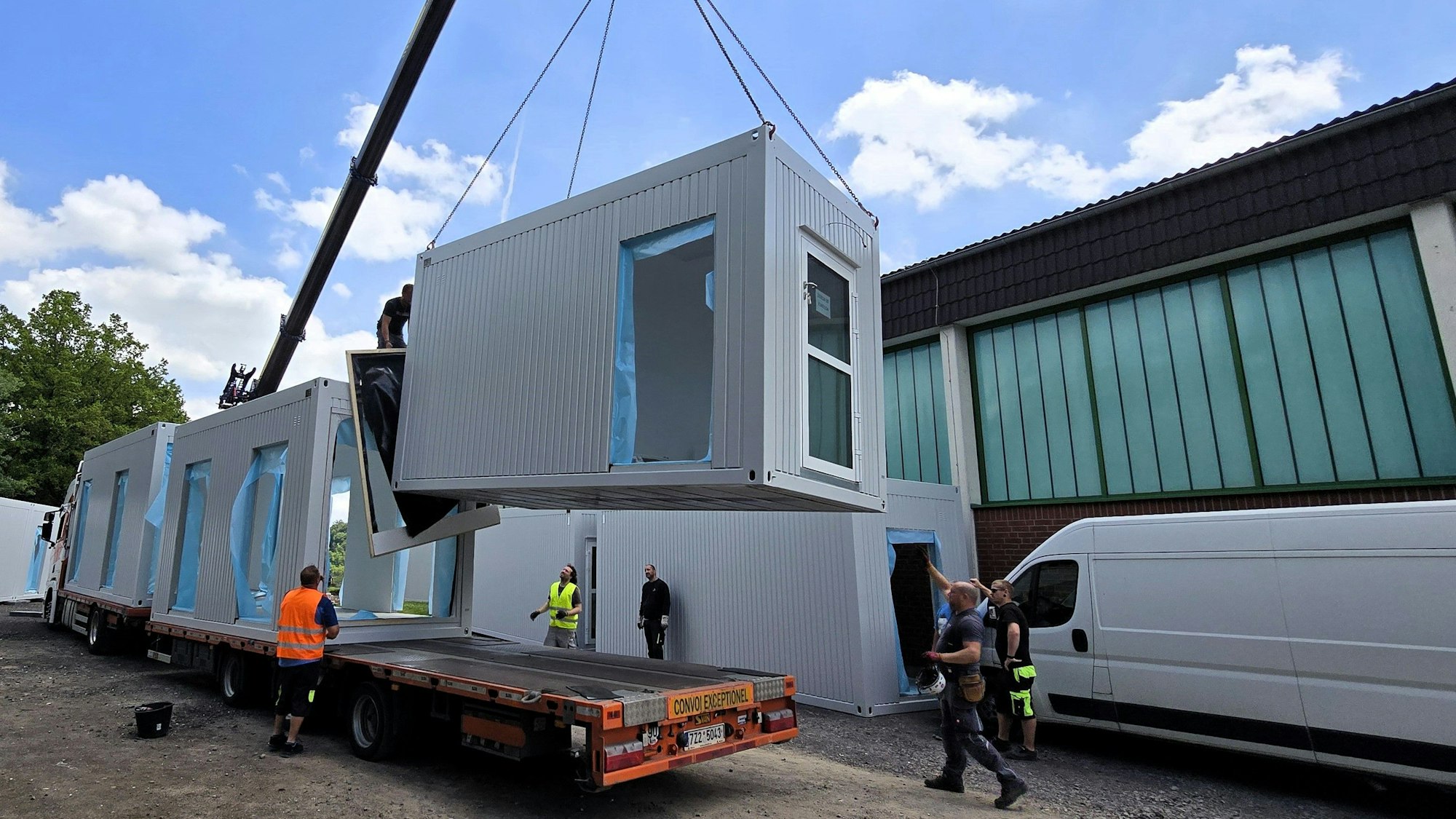 Ein Containermodul hängt am Haken eines Krans und wird von einem Auflieger gehoben.