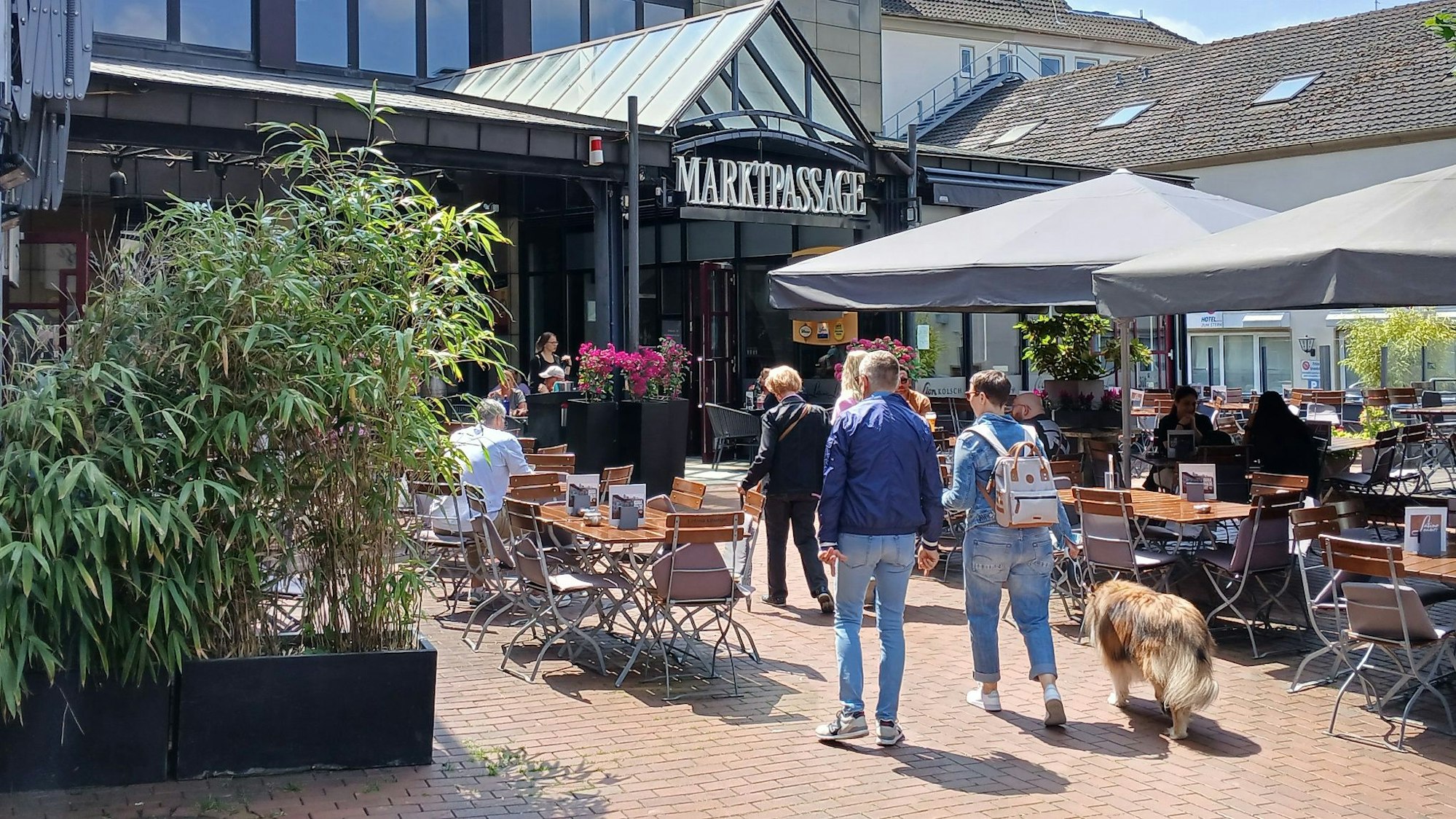 Menschen laufen zwischen den Tischen eines Außenlokals auf eine Einkaufspassage zu