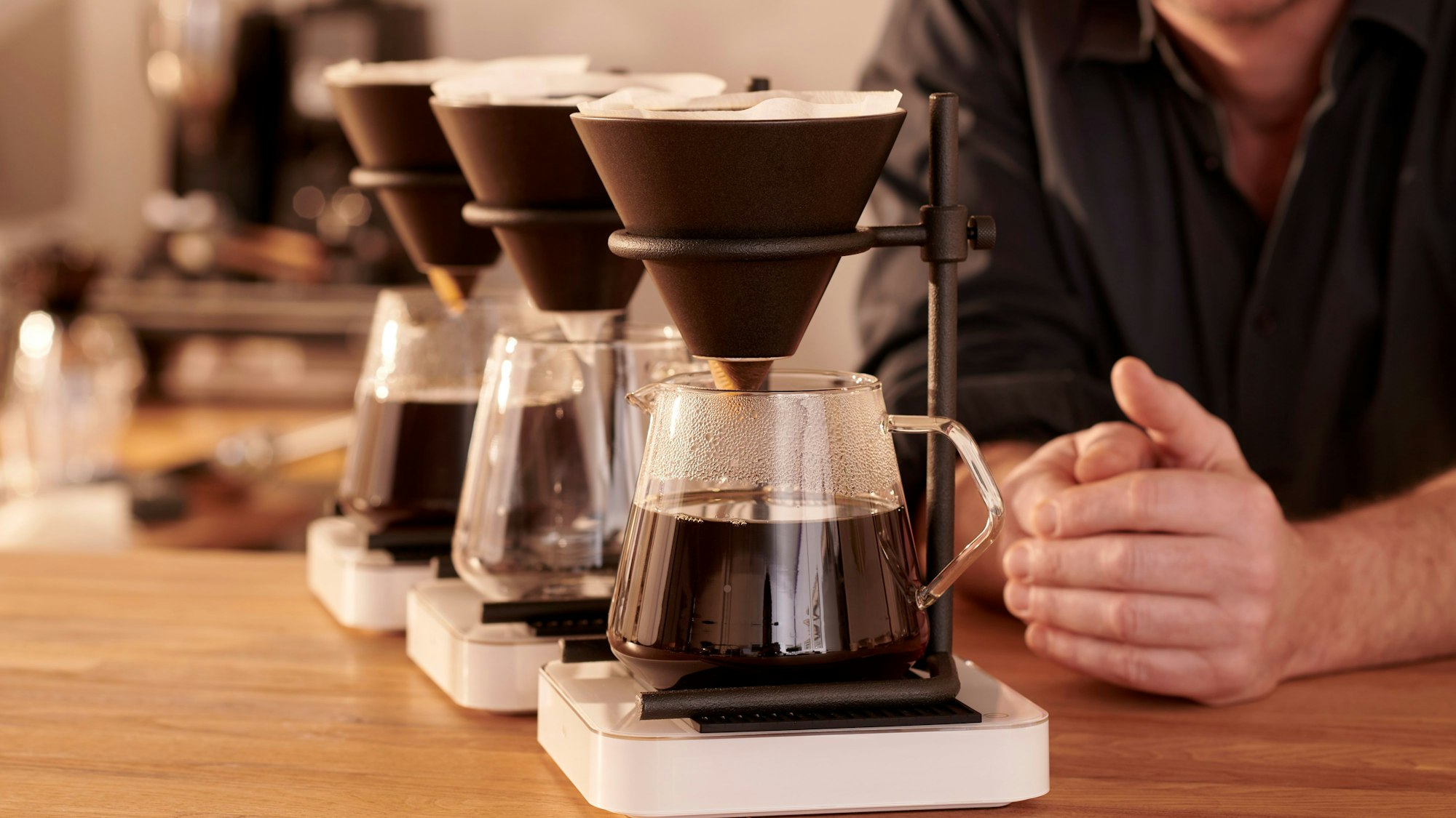 Mehrere Filterkannen mit Kaffee. Im Hintergrund sind die Hände einer Person zu sehen.