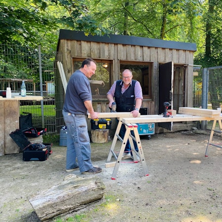 Zwei Männer sägen Holz vor einem Stall.