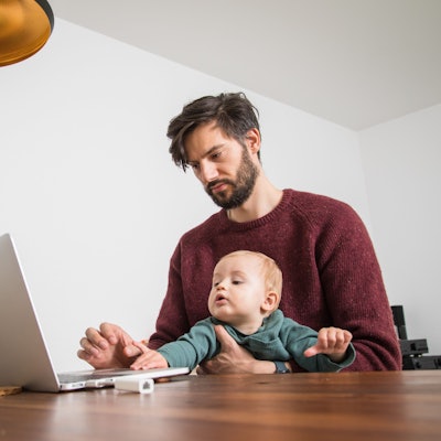 Ein Vater sitzt am Laptop, auf seinem Schoß ein Kleinkind, das sich Richtung Computer streckt.