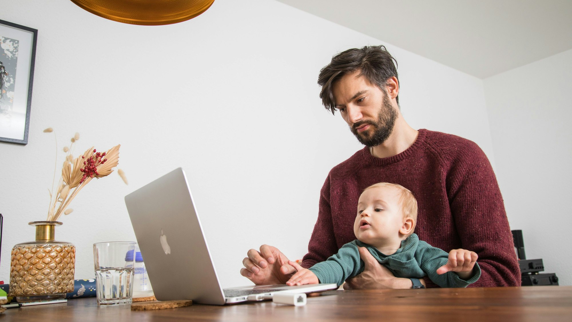 Ein Vater sitzt am Laptop, auf seinem Schoß ein Kleinkind, das sich Richtung Computer streckt.