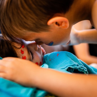 Zwei geschminkte Kinder stupsen ihre Nasen aneinander, eines der beiden liegt im Bett