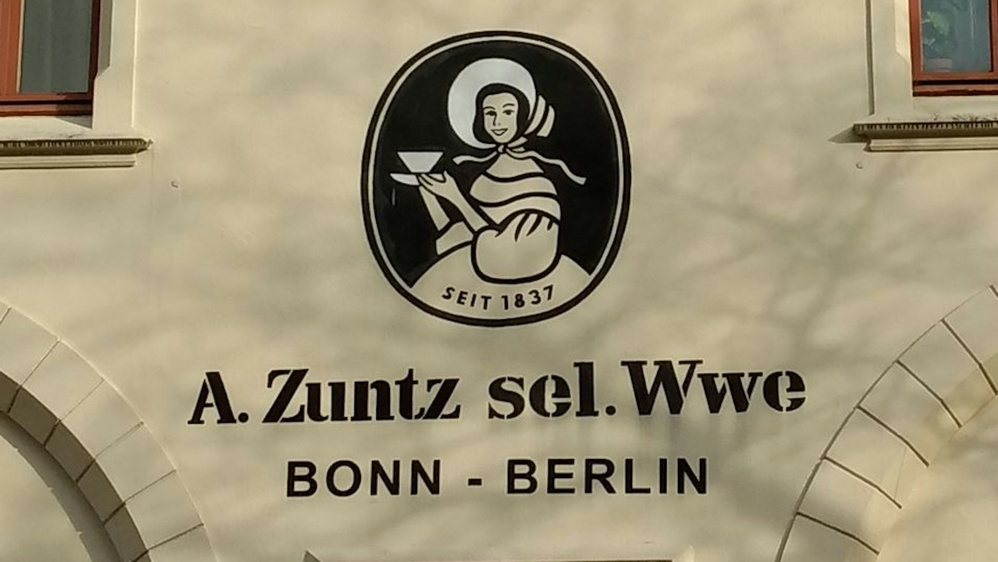 Das Logo einer Frau im Biedermeier-Kleid, mit einem Schutenhut und einer Kaffeetasse in der Hand. Darunter die Worte „A. Zuntz sel. Wwe“ und darunter „Bonn – Berlin“.