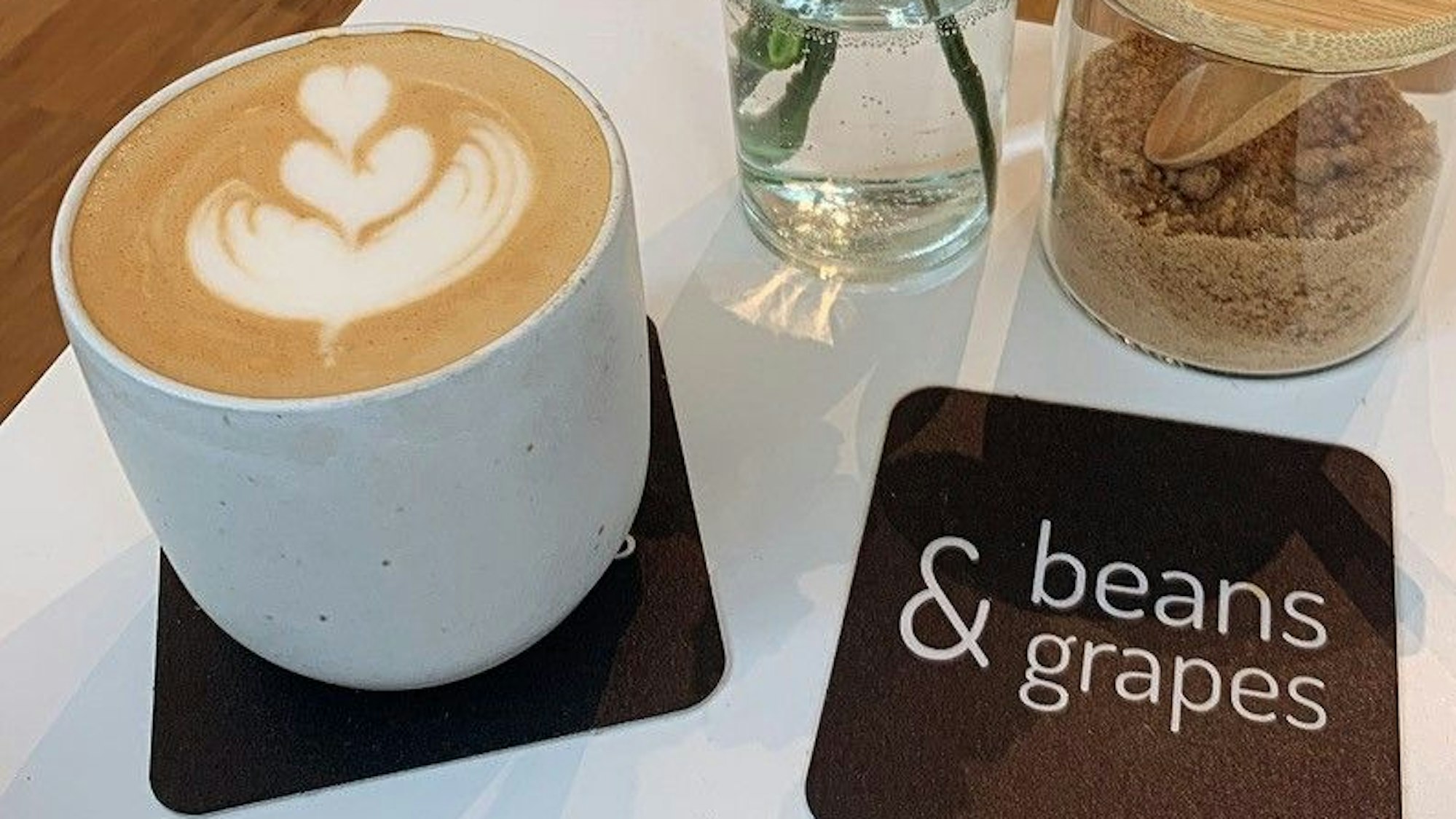 Ein Kaffee mit Muster auf Milchschaum, daneben ein Untersetzer mit der Aufschrift „Beans & Grapes“. Im Hintergrund brauner Zucker und eine Vase mit Blumen.
