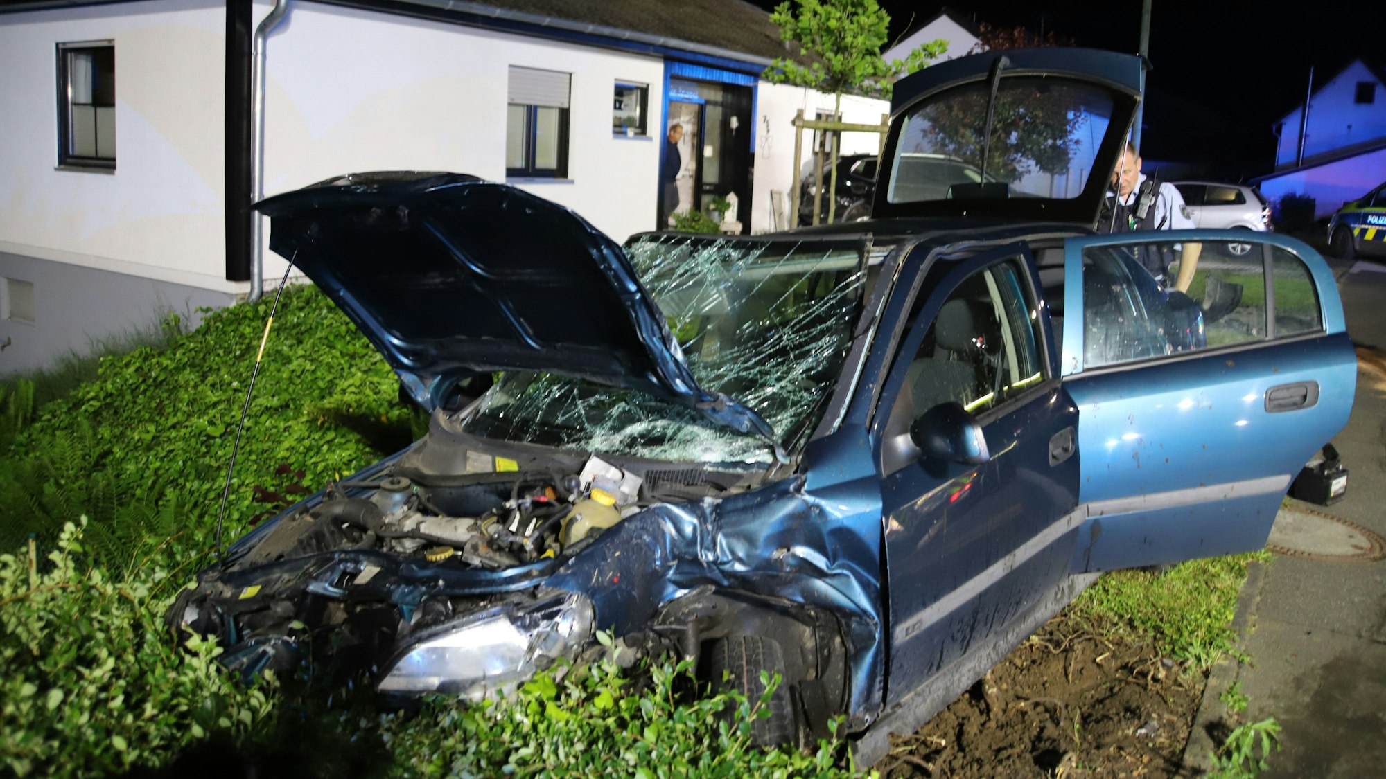 Ein demoliertes Auto steht in einer Hecke vor einem Haus.