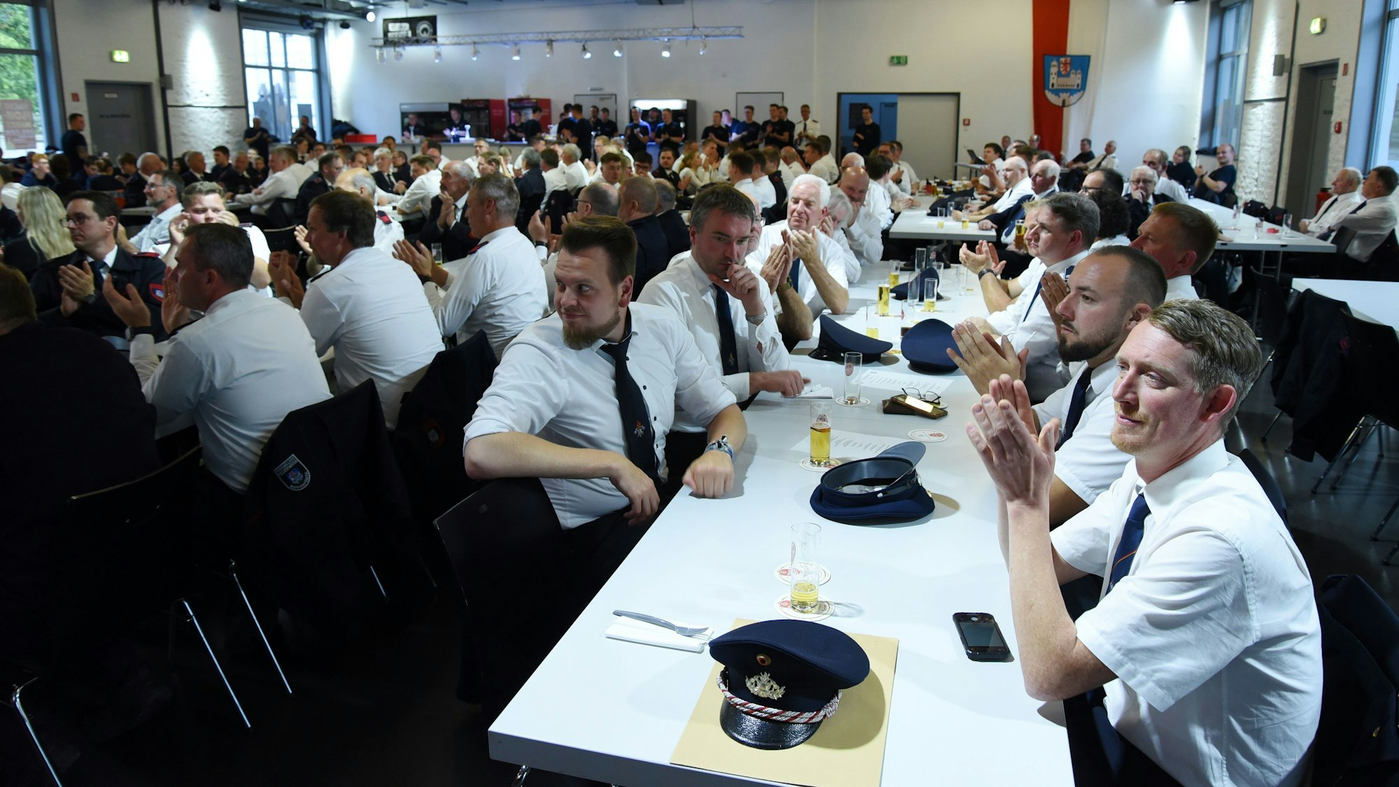 Zahlreiche Feuerwehrleute sitzen in einer Veranstaltungshalle an langen Tischreihen beisammen.