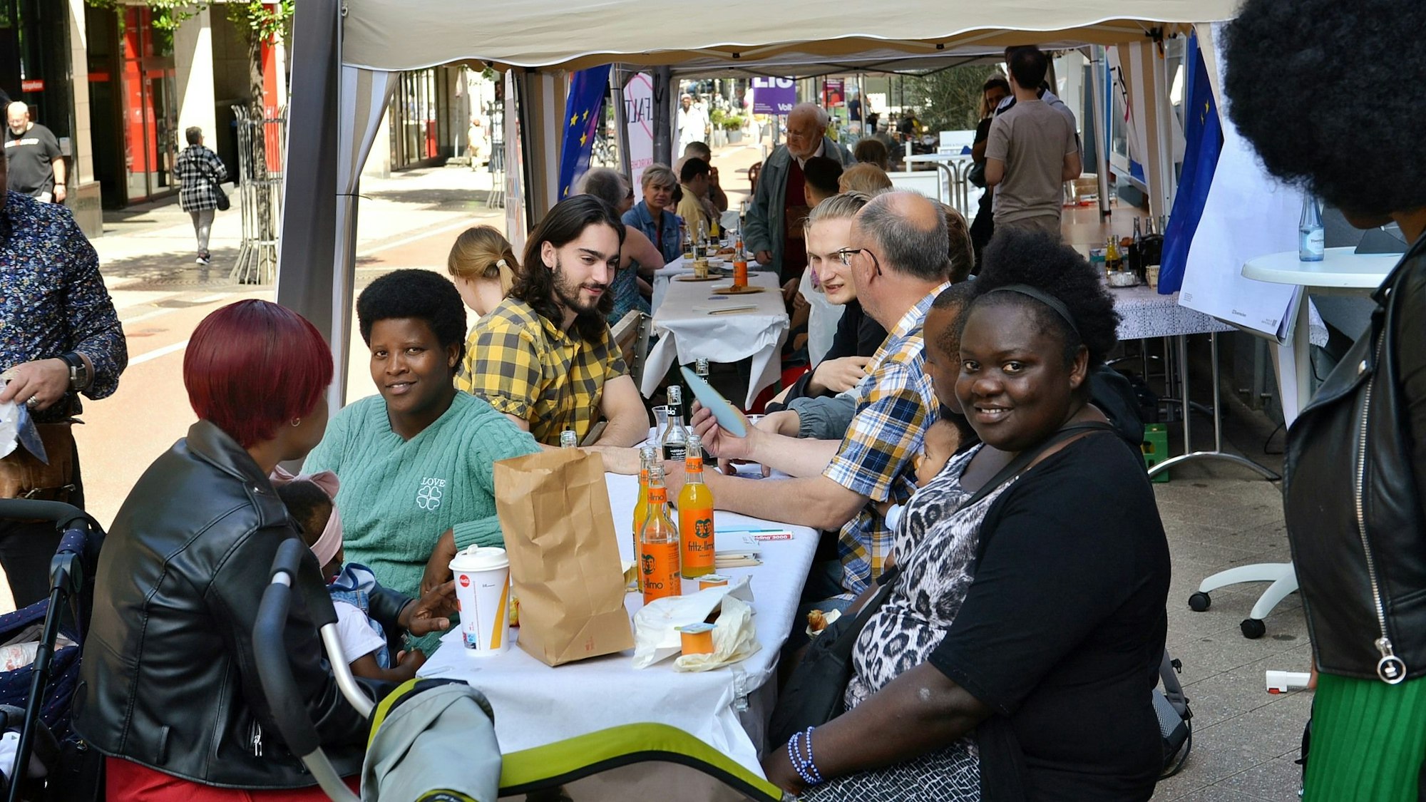 Das Bild zeigt Menschen unterschiedlichen Geschlechts und unterschiedlicher Hautfarbe, die an Tischen sitzen und sich unterhalten.