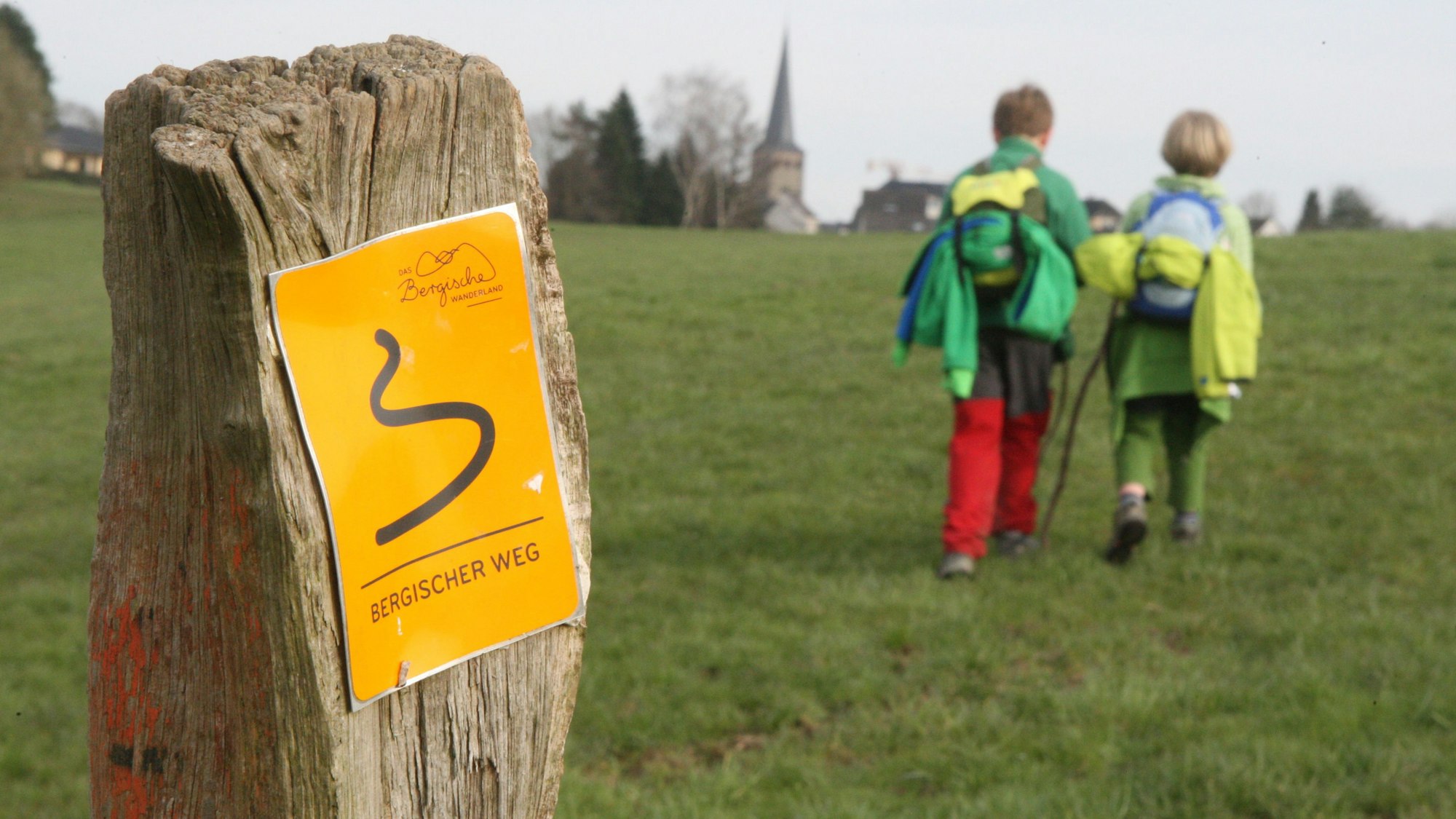 Zwei Kinder wandern auf dem Bergischen Weg in Richtung der Kirche von Neunkirchen.