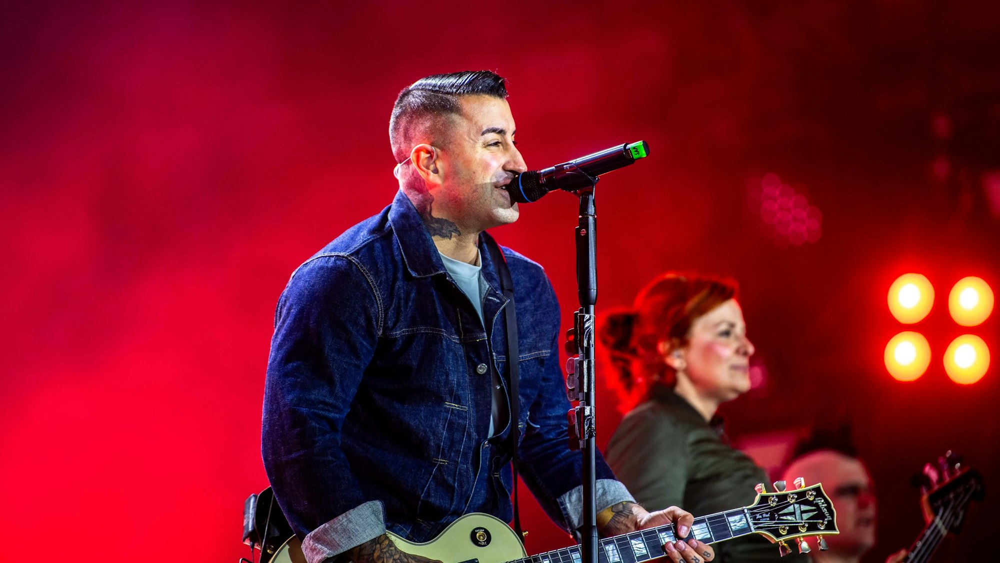 Sammy Amara steht in Jeansjacke an einem Mikrofon, singt und spielt Gitarre. Im Hintergrund ist Ines Maybaum zu erkennen. Die Bühne ist in rotes Licht getaucht.