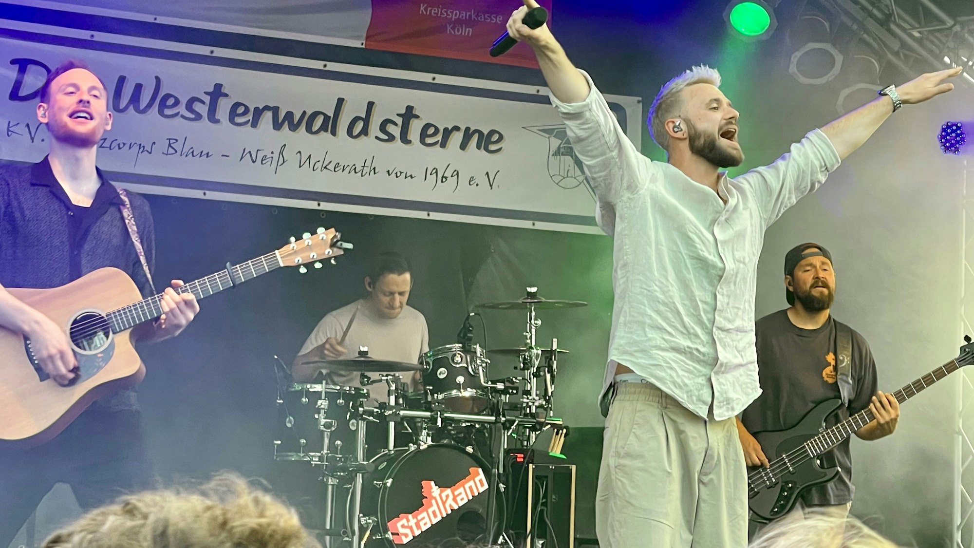 Eine Band steht auf der Bühne, der Sänger hat die Arme ausgebreitet.