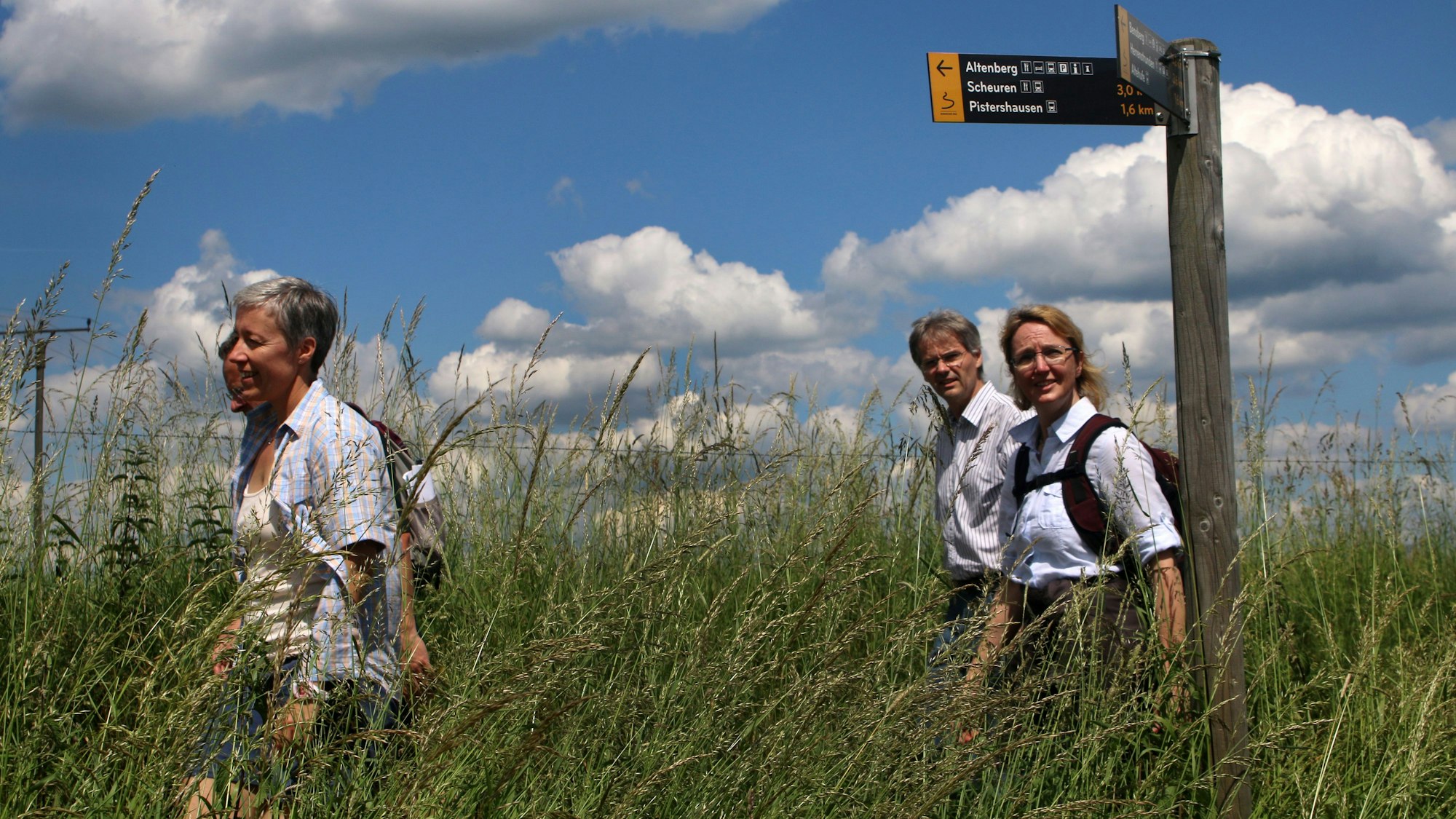 Menschen folgen einem Wegweiser des Bergischen Wegs bei Odenthal-Eikamp durch eine hohe Wiese.