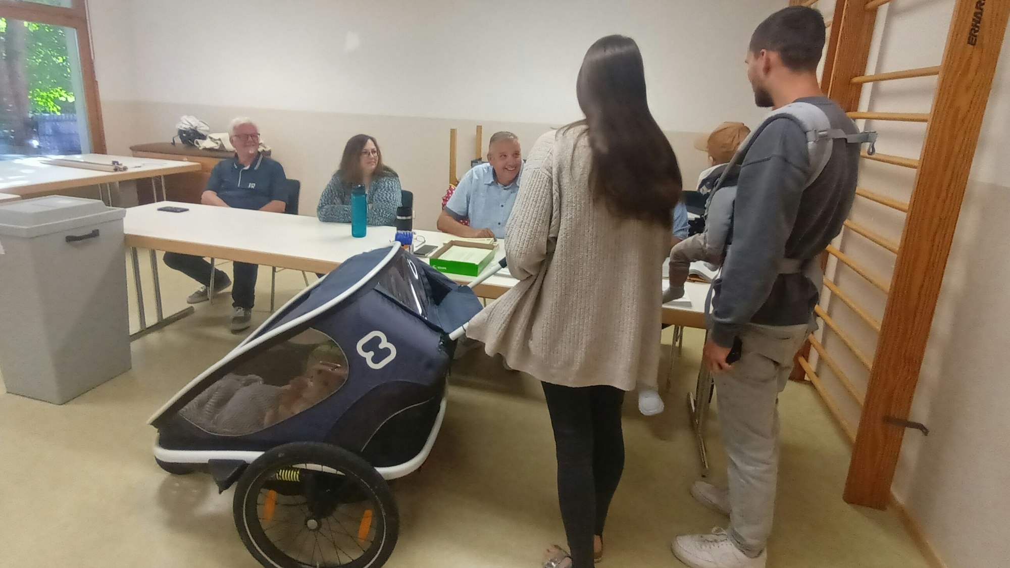 Vier Personen sitzen an einem Tisch, davor eine Familie mit Baby und Kinderwagen