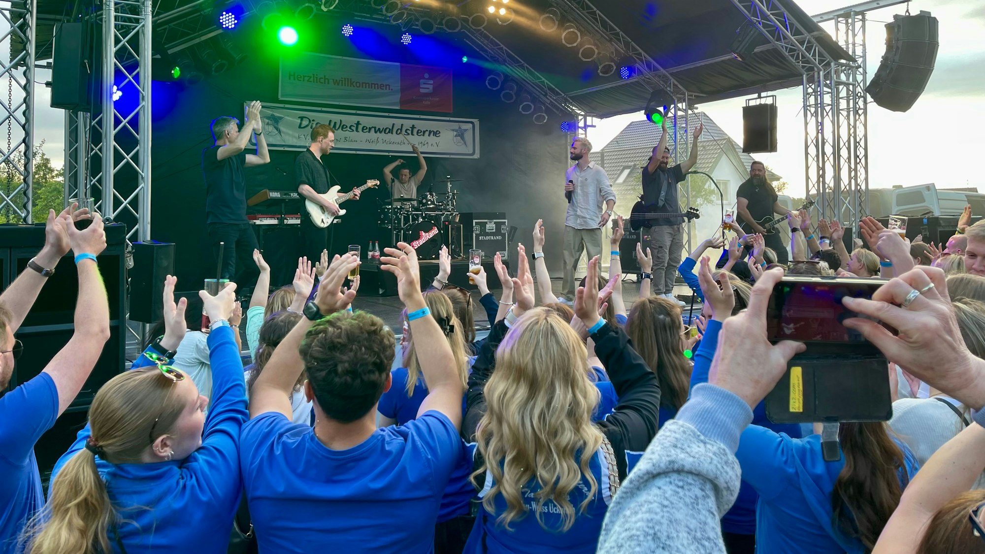 Dicht gedrängt steht Publikum vor einer Bühne, viele Menschen klatschen über ihren Köpfen in die Hände.