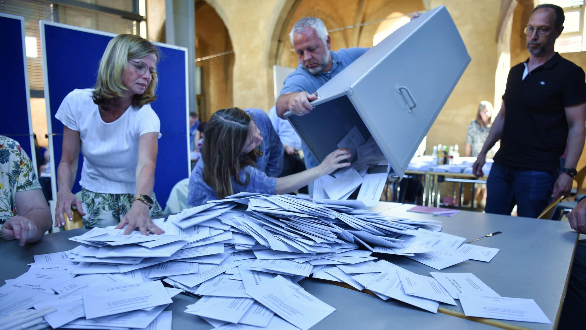 Eine Wahlurne wird von einem Mann geleert. Um den Tisch mit den Wahlzetteln stehen Wahlhelfer.