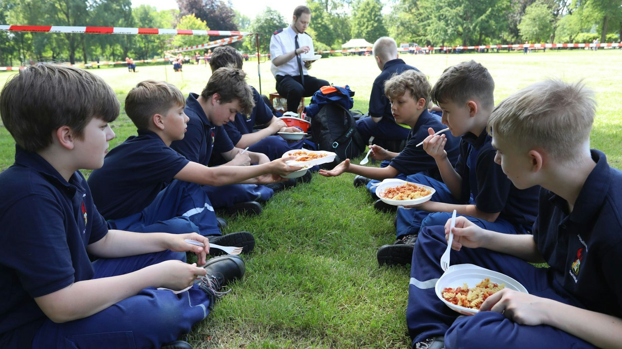 Jugendliche in blauer Uniform sitzen auf einer Wiese und essen Nudeln