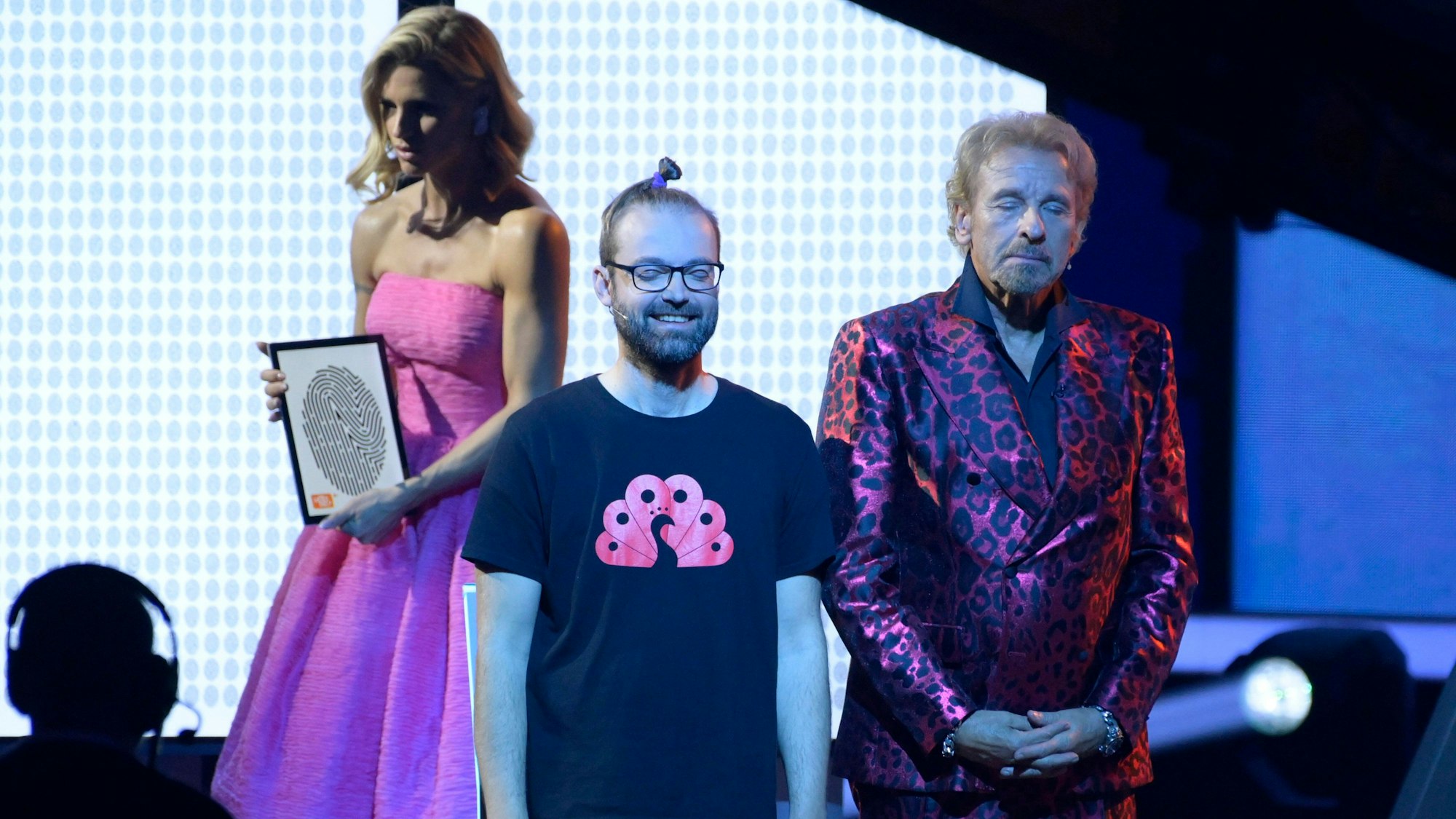 Eine Szene aus der TV-Show 2022: Reiß und Thomas Gottschalk sehen mit geschlossenen Augen auf der Bühne; Co-Moderatorin Michelle Hunziker hält ein Bild mit einem Fingerabdruck in die Kamera.