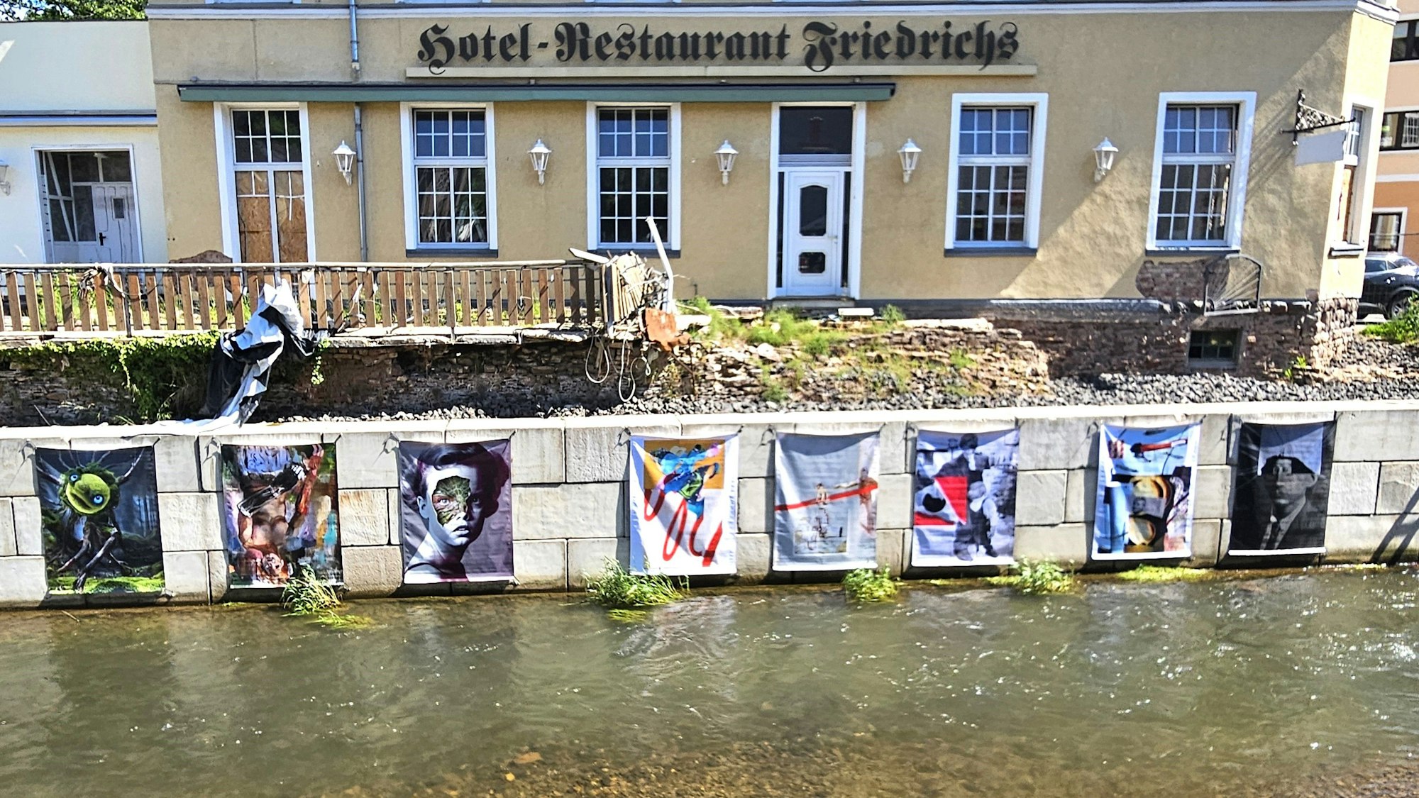 Die Schutzmauer an der Urft in Gemünd wurde mit großformatigen Kunstwerken bestückt, dahinter ist das von der Flut zerstörte Hotel Friedrichs zu sehen.