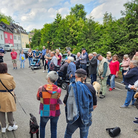 Bürgermeister Ingo Pfennings steht vor einem Haus in Gemünd und spricht in ein Mikrofon, die Teilnehmer des Spaziergangs stehen im Halbkreis vor ihm.